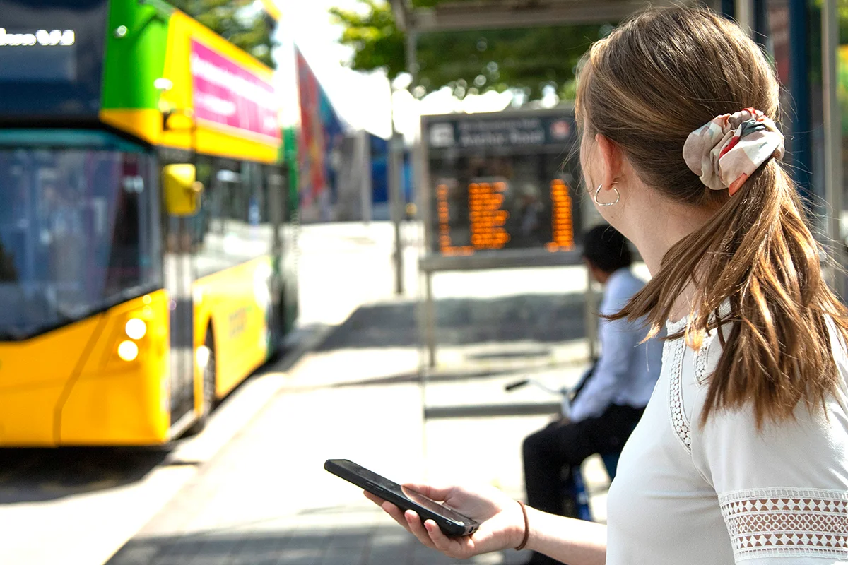 Community Get Moving young female waiting at bus stop with mobile in hand as bus arrives