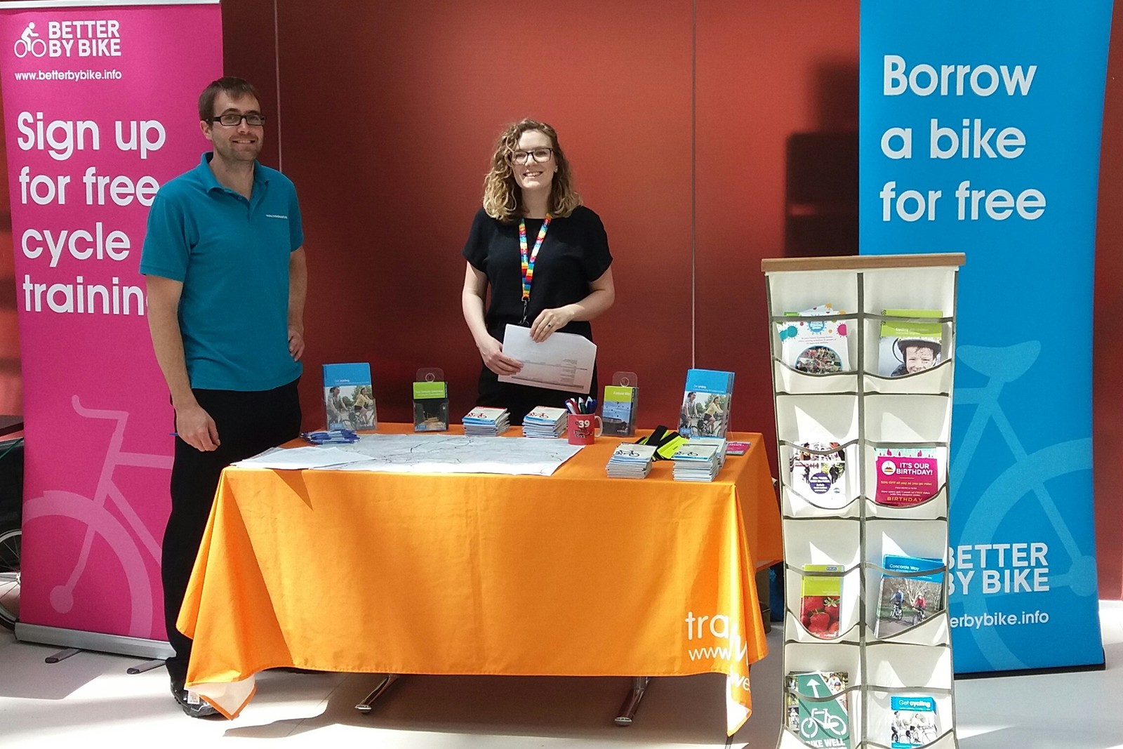 Two people behind a stand with colourful banners, posters and leaflets.